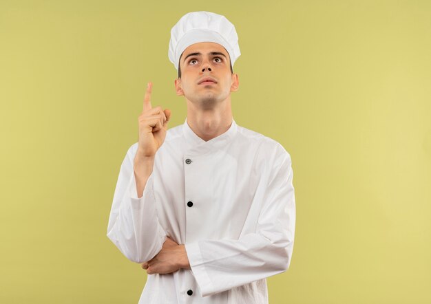 Mirando a un joven cocinero con uniforme de chef apunta con el dedo hacia arriba en la pared verde aislada con espacio de copia