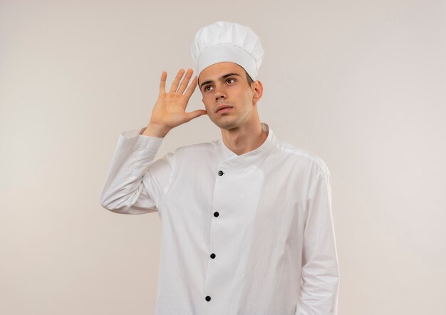 Mirando a un joven cocinero con uniforme de chef mostrando gesto de escucha en la pared blanca aislada con espacio de copia