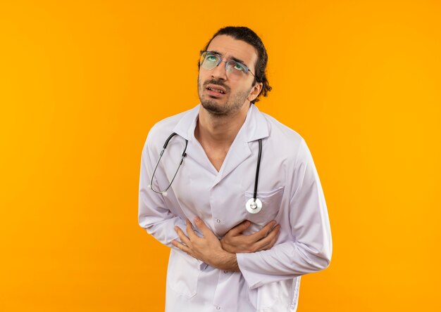 Mirando a joven médico enfermo con gafas médicas vistiendo bata médica con estetoscopio agarró el estómago