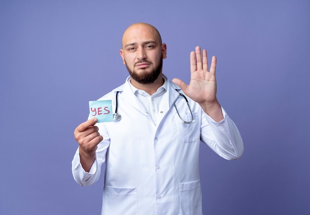 Mirando joven médico varón calvo vistiendo bata médica y estetoscopio sosteniendo la marca de papel y mostrando gesto de parada