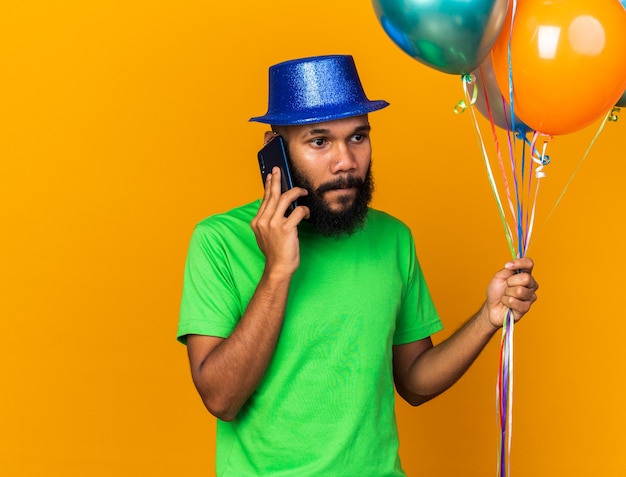 Mirando de lado joven afroamericano con gorro de fiesta sosteniendo globos habla por teléfono