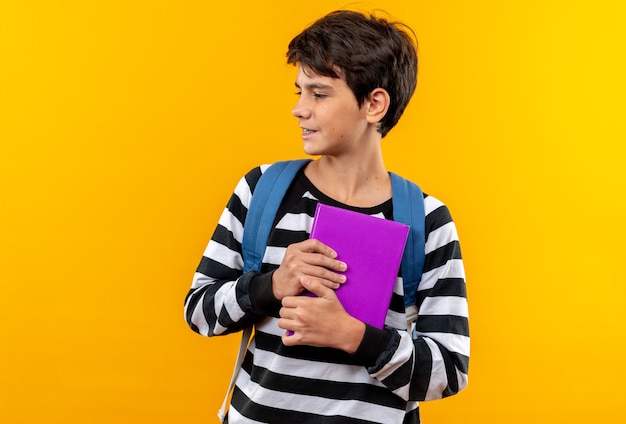 Mirando de lado joven escolar vistiendo mochila sosteniendo libro