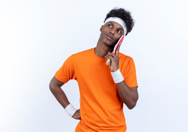 Mirando de lado pensando joven deportivo afroamericano vistiendo diadema y muñequera poniendo raqueta de ping pong en la mejilla aislado sobre fondo blanco.