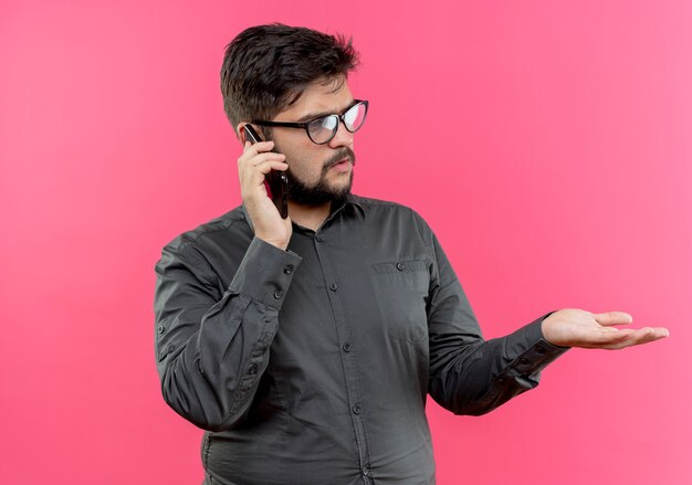 Mirando de lado pensando joven empresario con gafas habla por teléfono y extendiendo la mano al lado aislado en la pared rosa