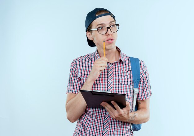 Mirando de lado pensando joven estudiante niño con mochila y gafas y gorra sosteniendo el portapapeles y manteniendo la pluma en la mejilla