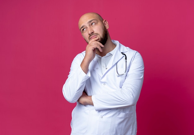 Mirando de lado pensando joven médico varón calvo vistiendo bata médica y estetoscopio poniendo la mano en el mentón aislado sobre fondo rosa con espacio de copia