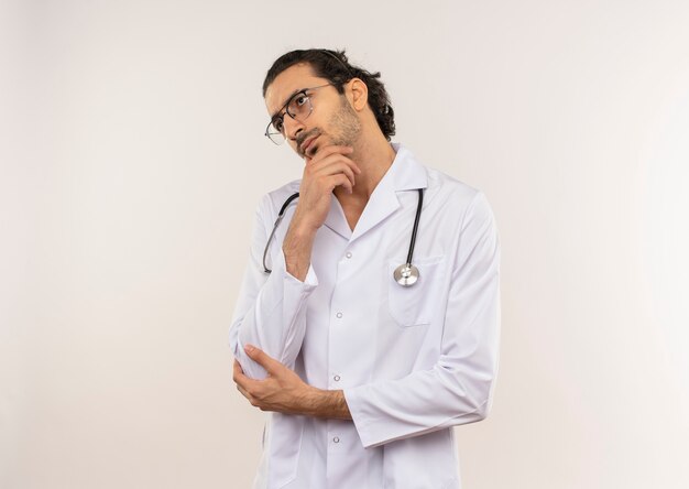 Mirando de lado pensando joven médico varón con gafas ópticas vistiendo túnica blanca con estetoscopio poniendo la mano en la barbilla