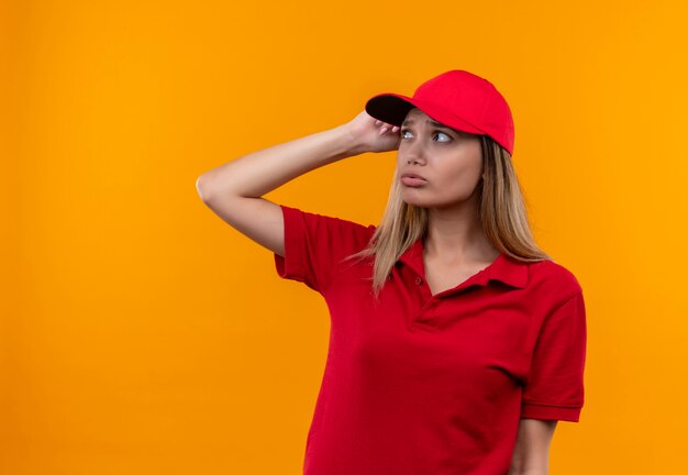 Mirando de lado pensando joven repartidora vestida con uniforme rojo y gorra poniendo la mano en la cabeza