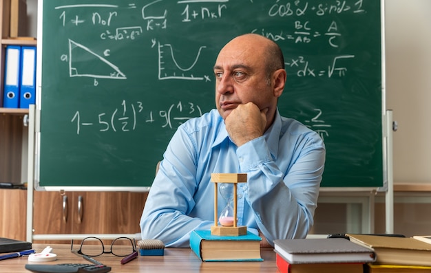 Mirando de lado el profesor de mediana edad se sienta a la mesa con útiles escolares en el aula