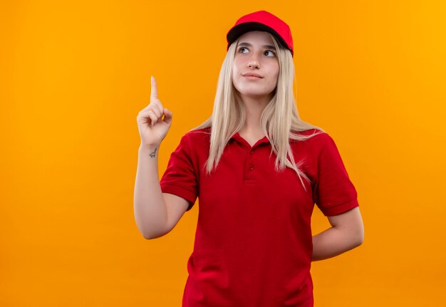 Mirando a la mujer joven de entrega lateral con camiseta roja y gorra apunta hacia arriba en la pared naranja aislada