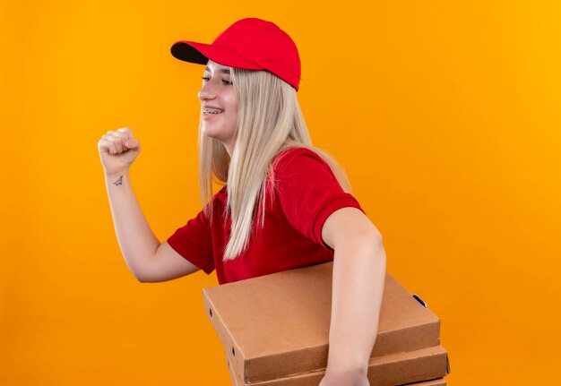 Mirando a la mujer joven de entrega lateral con camiseta roja y gorra en corsé dental sosteniendo una caja de pizza que muestra un gesto de sí en la pared naranja aislada