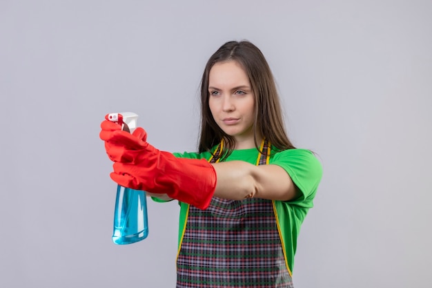 Mirando a la mujer joven de limpieza lateral con uniforme en guantes rojos sosteniendo spray de limpieza a un lado en la pared blanca aislada