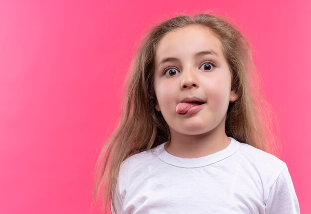 Mirando a la niña de la escuela vistiendo camiseta blanca mostrando lengua sobre fondo rosa aislado