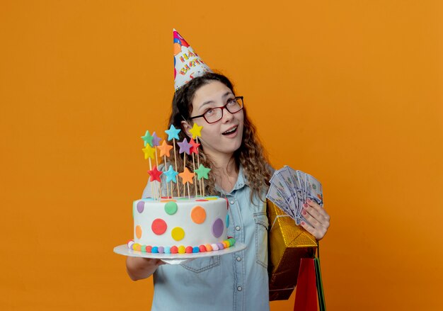 Mirando a la niña con gafas y gorro de cumpleaños sosteniendo pastel de cumpleaños con bolsas de regalo con cajas y dinero aislado sobre fondo naranja