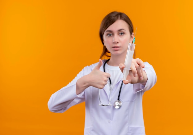 Mirando seriamente a la joven doctora vistiendo una bata médica y un estetoscopio estirando la jeringa y mostrando el pulgar hacia arriba en el espacio naranja aislado con espacio de copia