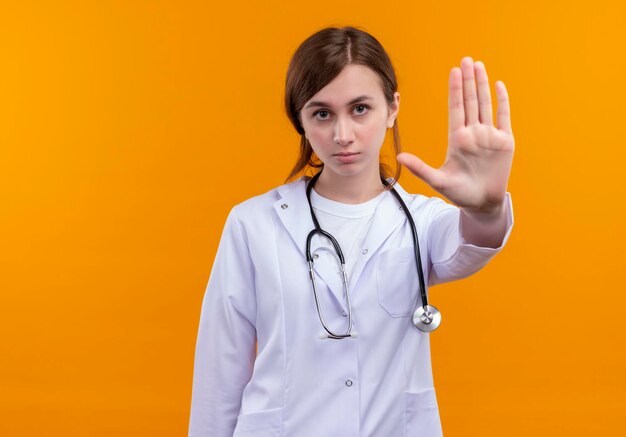 Mirando seriamente a la joven doctora vistiendo una bata médica y un estetoscopio haciendo gesto de parada en un espacio naranja aislado con espacio de copia
