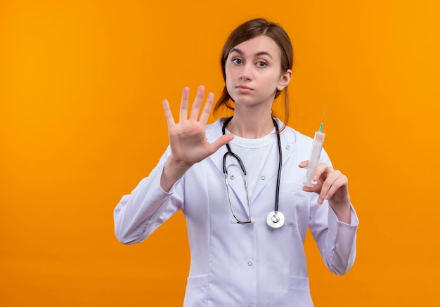 Mirando seriamente a la joven doctora vistiendo bata médica y estetoscopio sosteniendo una jeringa y mostrando cinco en un espacio naranja aislado con espacio de copia