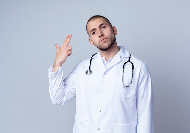 Mirando seriamente a joven médico vistiendo bata médica y estetoscopio alrededor de su cuello haciendo gesto de pistola con la mano aislada en la pared blanca