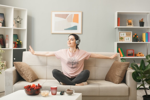 Mirando sonriente joven de lado usando audífonos haciendo yoga sentado en el sofá detrás de la mesa de café en la sala de estar