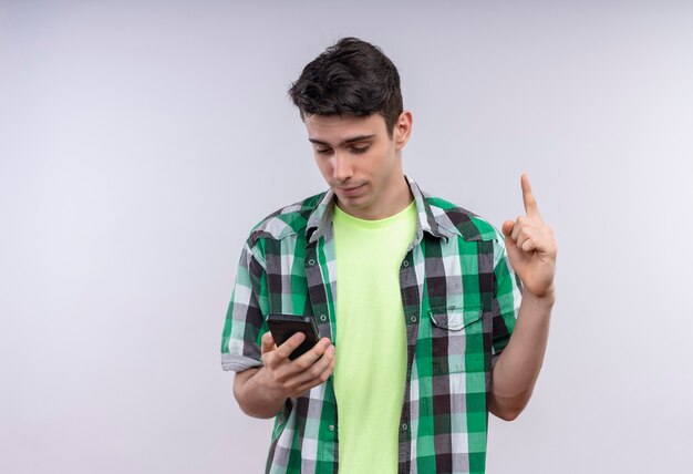 Mirando el teléfono joven caucásico con camisa verde apunta hacia arriba en la pared blanca aislada