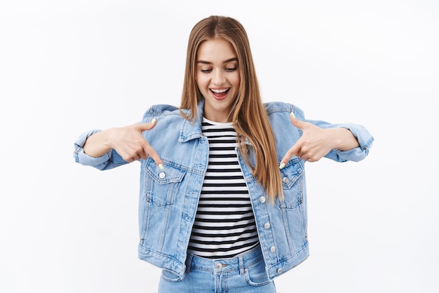 Foto gratuita mire aquí retrato de niña rubia sonriente mirando hacia abajo apuntando hacia abajo anuncio ver producto genial evento impresionante promoción festival de música juvenil de pie fondo blanco