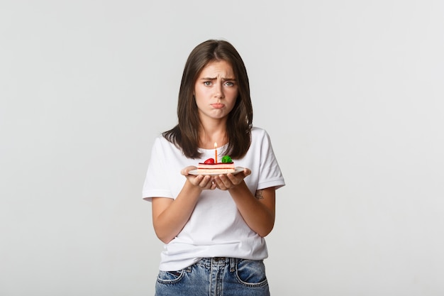 Foto gratuita miserable cumpleañera con cara de tristeza, sosteniendo b-day cake con vela.