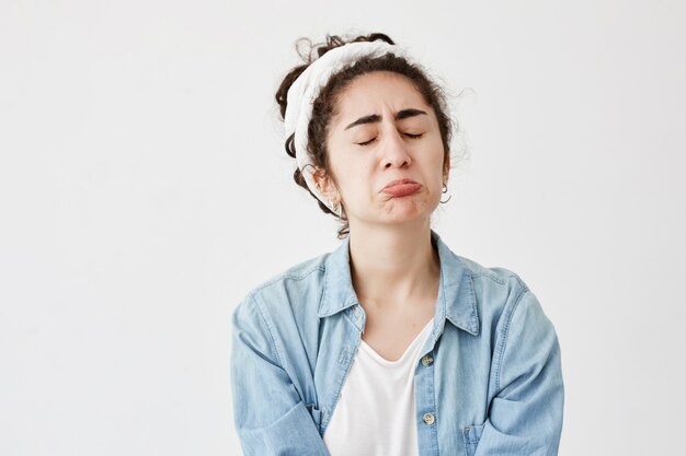 Miserable mujer triste y triste con cabello oscuro y ondulado se ve ofendida, frunce los labios, frunce el ceño con frustración, frustrada, después de recibir malas noticias. Infeliz joven en camisa vaquera llora de frustración