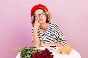 Foto gratuita miserable ofendida insatisfecha linda chica curva labios sentado solo junto a la mesa con café y croissant en rosa.