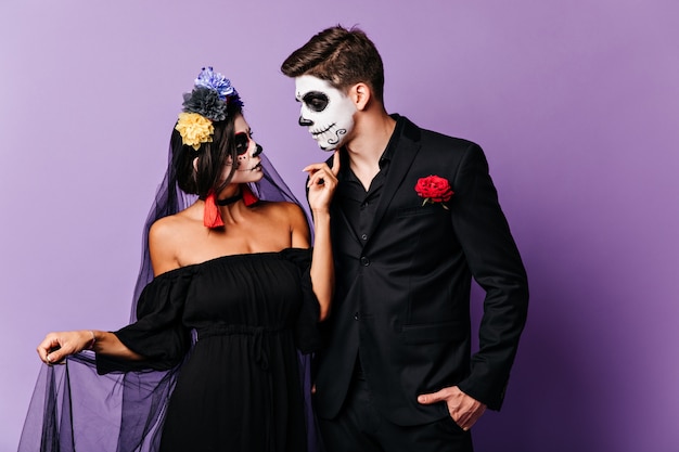 Misteriosa chica con la cara pintada acariciando suavemente a su amado novio. Foto de pareja en trajes negros con acentos brillantes posando sobre fondo morado.