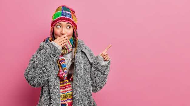 misteriosa joven asiática cubre la boca con la mano dice que el secreto indica que se encuentra en el espacio de la copia viste un abrigo de piel gris bufanda tejida y un sombrero aislado sobre una pared rosa