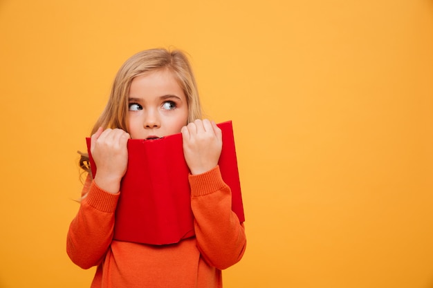 Misteriosa jovencita en suéter escondiéndose detrás del libro y mirando a otro lado sobre naranja