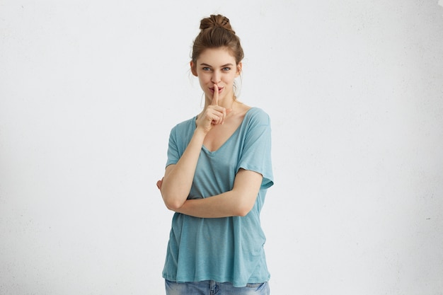 Misteriosa mujer bonita con moño de pelo con camiseta casual azul suelta sosteniendo el dedo índice en los labios con aspecto enigmático. Hermosa mujer con ojos azules encantadores pidiendo mantener el secreto y estar en silencio