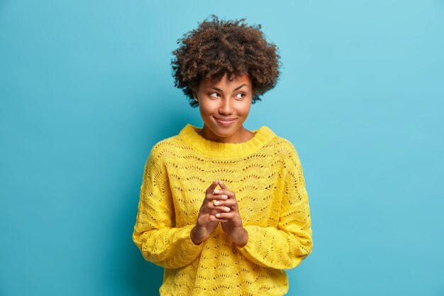 Misteriosa mujer se frota las manos y planea algo que los dedos de los empinados tienen un gran plan en mente vestida con un jersey amarillo de punto aislado sobre una pared azul insinúa con una sonrisa