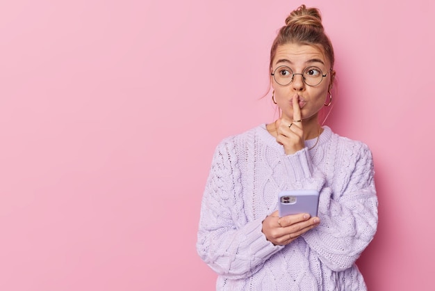 Foto gratuita misteriosa mujer joven y bonita muestra gesto de silencio exige silencio sostiene el teléfono móvil envía mensajes de texto usa gafas redondas y suéter de punto aislado sobre un espacio de maqueta de fondo rosa