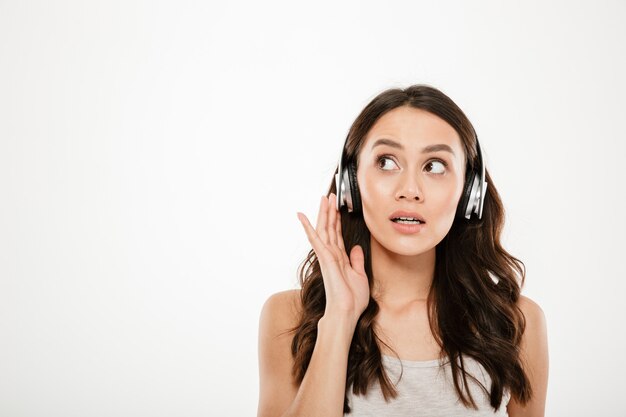 Misteriosa mujer morena en auriculares escuchando música y mirando a otro lado sobre gris
