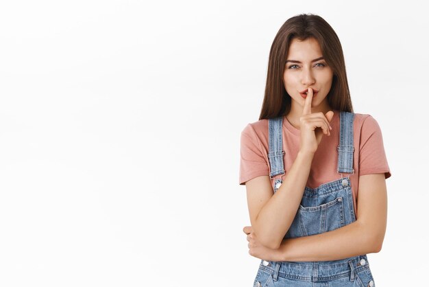 Misteriosa y seductora mujer morena moderna con camiseta de mono prepara una sorpresa pidiendo que guardes silencio sobre su secreto callando y diciendo shhh mientras presionas el dedo índice en los labios entrecerrando los ojos astuto fondo blanco