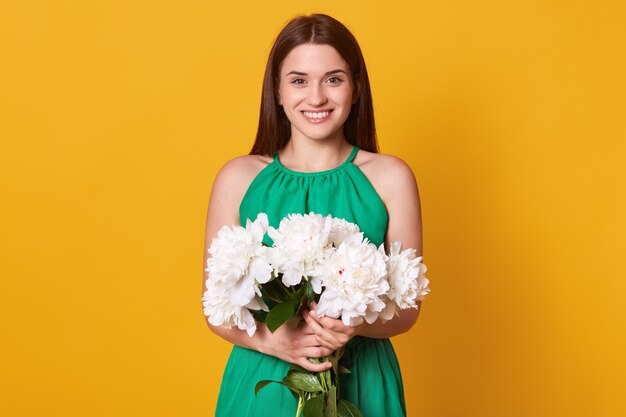 La mitad de la dama con un elegante vestido verde mantiene el ramo de flores en las manos en amarillo, feliz de recibir peonías como regalo.