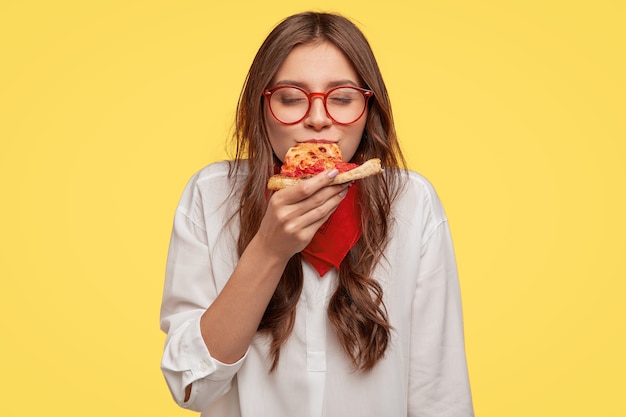 Mmm, ¡qué delicioso! Una mujer bonita de cabello oscuro come una rebanada de pizza italiana, mantiene los ojos cerrados por el placer, disfruta del buen gusto, usa gafas y camisa, aislada sobre una pared amarilla. Concepto de alimentación