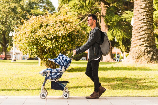 Mochila que lleva del hombre elegante que camina con el cochecito de bebé en el parque