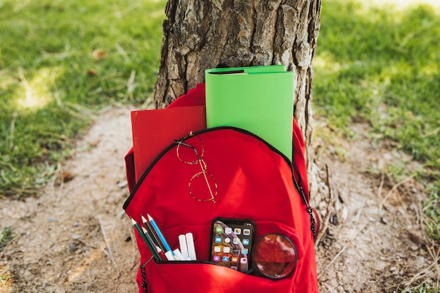 Mochila roja con papelería y teléfono inteligente cerca del árbol.