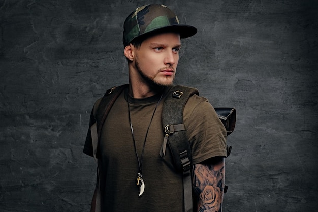 Mochilero de estilo urbano con brazos tatuados, vestido con camiseta verde oscuro y gorra Camo.