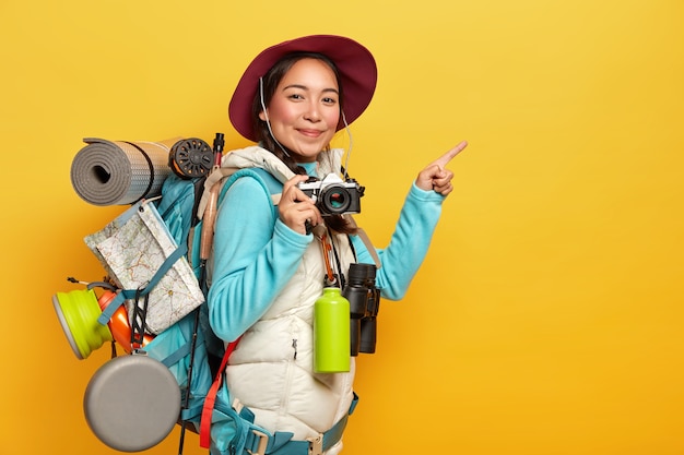 Mochilero femenino activo señala con el dedo índice en el espacio de la copia a un lado, sostiene una cámara retro, toma fotografías, lleva mochila, binoculares y termo, usa ropa informal