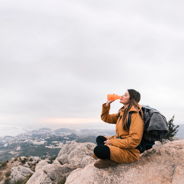 Mochilero femenino sentado en la cima de la montaña bebiendo el agua