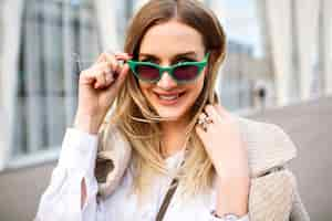 Foto gratuita moda al aire libre de cerca el retrato de la impresionante mujer rubia de negocios, sonriendo y mirando a la cámara, abrigo de cachemira, gafas de sol de ojo de gato vintage, joyas, colores suaves.