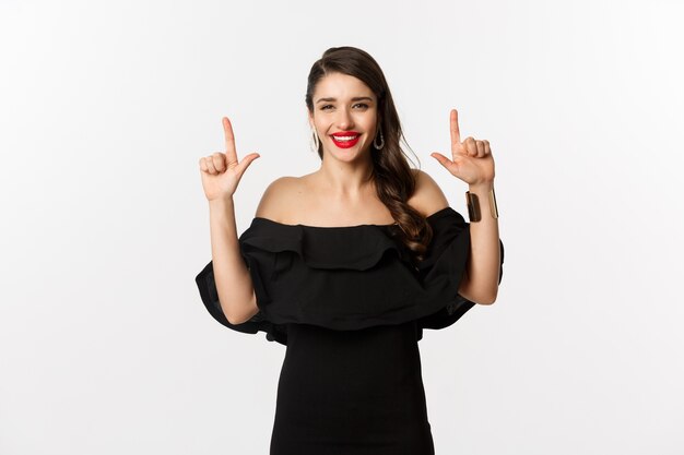 Moda y belleza. Encantadora mujer con labios rojos, vestido negro, sonriendo feliz y señalando con el dedo hacia arriba, mostrando el logo, fondo blanco.