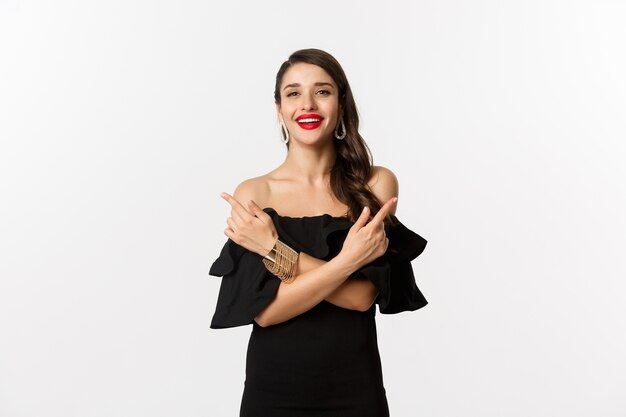 Moda y belleza. Hermosa mujer feliz apuntando hacia los lados, mostrando dos opciones y sonriendo, vestida con un vestido negro, fondo blanco.