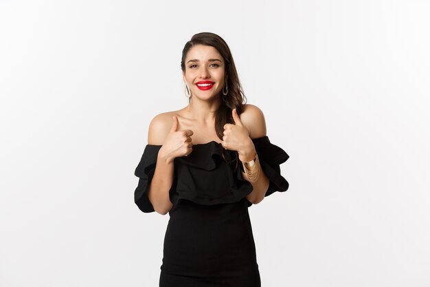 Moda y belleza. Hermosa mujer vestida de negro, lápiz labial rojo, mostrando los pulgares hacia arriba en señal de aprobación, recomendando el producto, de pie sobre fondo blanco.