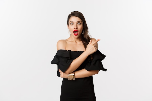 Moda y belleza. Hermosa mujer en vestido negro, labios rojos, apuntando con el dedo a la derecha en la oferta promocional, mirando asombrado a la cámara, fondo blanco.