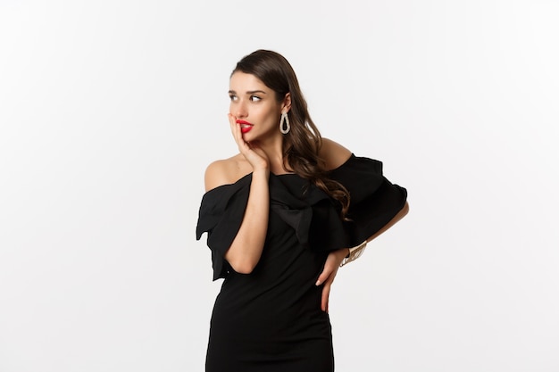 Moda y belleza. Imagen de elegante mujer hermosa en vestido negro y maquillaje, mirando a la izquierda con tentación, tocando labios rojos, de pie sobre fondo blanco.