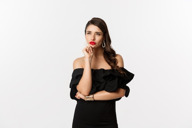 Moda y belleza. Imagen de mujer glamour pensativa mirando a otro lado, entrecerrando los ojos mientras piensa, de pie en vestido negro sobre fondo blanco.